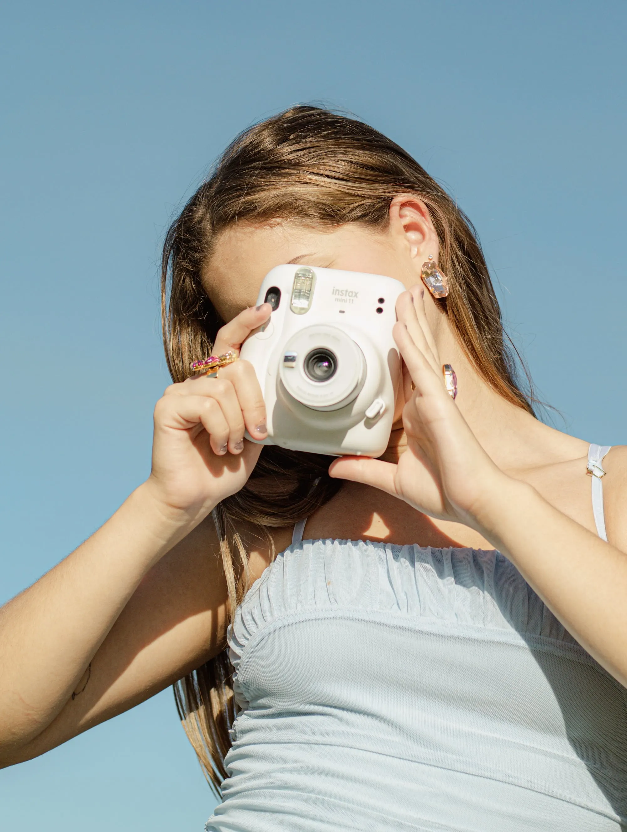 Modelo fotografando com uma câmera instantânea, usando brincos e anéis com pedras coloridas